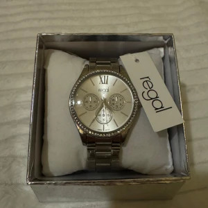 Silverfärgad klocka från Regal - Elegant silverfärgad klocka från Regal med glittrande stenar runt urtavlan. Klockan har en stilren design med romerska siffror och tre mindre urtavlor för extra funktionalitet. Perfekt accessoar för att lyfta din stil.