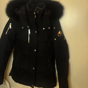 Svart dunjacka från Moose Knuckles - Säljer en svart dunjacka från Moose Knuckles med pälskantad huva och flera dragkedjefickor. Jackan är äkta och kvitto finns. Använd ett fåtal gånger. 