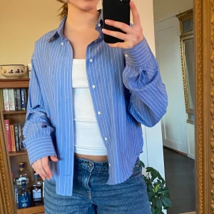 Blå randig skjorta - Snygg blå skjorta med vita ränder och knappar. Skjortan har en avslappnad passform och långa ärmar, perfekt för en stilren look.