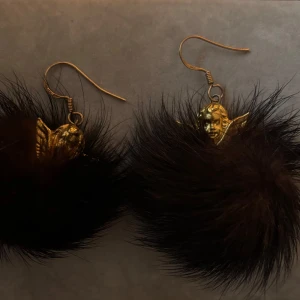 Guldörhängen med svarta/mörkbruna pälsbollar - Unika örhängen i guld med dekorativa svarta/mörkbruna pälsbollar. Perfekta för att ge en elegant touch till din stil. Hängande design med detaljerade guldfärgade änglar. 