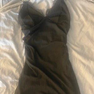 Svart klänning med spetsdetaljer - Elegant svart klänning med spetsdetaljer vid halsringningen. Klänningen har en omlott-design och smala axelband, vilket ger en stilren look. Perfekt för en kväll ute.