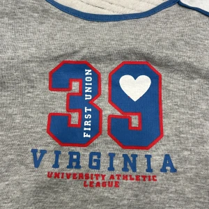 Grå t-shirt med tryck - Snygg grå t-shirt med tryck i blått och rött. Texten 'Virginia University Athletic League' och siffran '39' med ett hjärta ger en sportig känsla. Perfekt för en avslappnad stil.