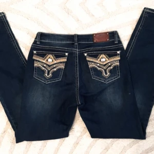 Mörkblå jeans med broderade fickor från Stars & Stripes - Köpf I Stockholm. Snygga mörkblå jeans med unika broderade detaljer och pärlor på bakfickorna. Dessa jeans har en klassisk femficksdesign och en knappgylf. Perfekta för dig som vill sticka ut med stil.