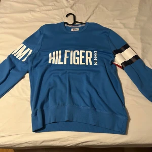 Blå tröja från Hilfiger Denim - Snygg blå tröja från Hilfiger Denim med stora vita bokstäver på bröstet och ärmarna. Tröjan har långa ärmar och en klassisk rund halsringning. Perfekt för en avslappnad stil.
