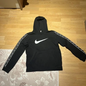 Svart hoodie från Nike - Säljer en svart hoodie från Nike med det klassiska vita swoosh-logotypen på bröstet. Tröjan har långa ärmar med vita detaljer längs ärmarna och en praktisk magficka. Perfekt för en sportig look!