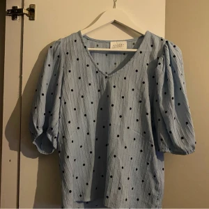 Blå blus med prickar - Charmig ljusblå blus med svarta prickar och v-ringning. Blusen har trekvartsärmar och en luftig passform, perfekt för vår och sommar. Den är tillverkad i ett lätt och bekvämt material.