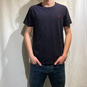 Marinblå t-shirt från Ralph Lauren - Snygg marinblå t-shirt från Ralph Lauren med det klassiska röda logotypbroderiet på bröstet. Perfekt för en stilren och avslappnad look. Storlek L men passar bättre på M, modellen är 180cm. 