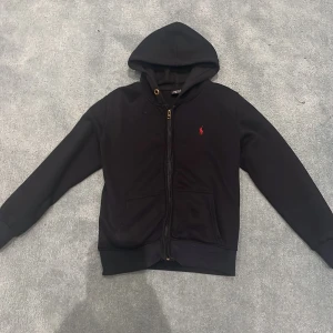 Mörkblå zip-up från Ralph Lauren - Säljer en klassisk mörkblå zip-up från Ralph Lauren med dragkedja och huva. Den har en liten röd logga på bröstet och är perfekt för en avslappnad stil. Passar bra till både jeans och träningsbyxor. 