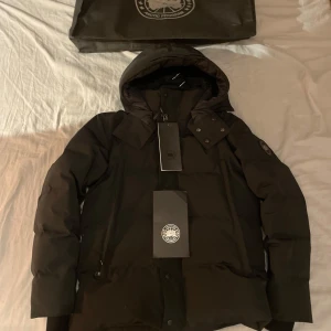 Svart Canada Goose black labal - Snygg och varm svart dunjacka från Canada Goose med huva och dragkedja. Jackan har långa ärmar och knappar framtill. Perfekt för kalla dagar.