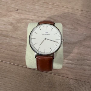 Klocka från Daniel Wellington - Snygg klocka från Daniel Wellington med en stilren vit urtavla och brunt skinnarmband. Klockan har en silverfärgad boett och minimalistisk design, perfekt för en elegant look. Klockan är knappt använd. Nypris ligger på cirka 2000kr