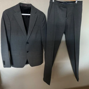 Grå kostym från Oscar Jacobson - Snygg grå kostym från Oscar Jacobson, perfekt för formella tillfällen. Kavajen har två knappar och byxorna har en klassisk passform. Tillverkad i en blandning av ull och polyester för en bekväm och stilren look.