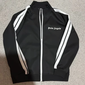 Svart track jacket från Palm Angels - Snygg svart track jacket från Palm Angels med vita ränder längs ärmarna och dragkedja framtill. Jackan har en hög krage och Palm Angels-logga på bröstet. Perfekt för en sportig look.