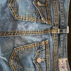 True religion jeans - Säljer ett par blå jeans med tydliga kontrastsömmar och unika fickdetaljer. Jeansen har en bootcut-stil och är perfekta för en avslappnad look. De har en klassisk design med knappar och dragkedja framtill.