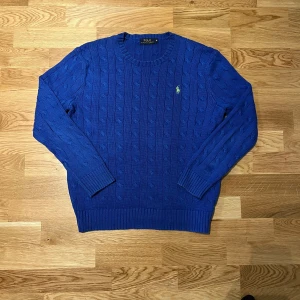 Blå stickad tröja från Ralph Lauren - Säljer en snygg blå stickad tröja från Ralph Lauren med kabelstickat mönster och det klassiska logotypbroderiet i grönt på bröstet. Tröjan har långa ärmar och ribbade muddar vid ärmslut och nederkant. Perfekt för kyligare dagar!