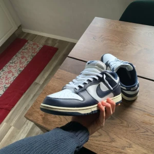 Nike Dunk i blått och vitt - Snygga Nike Dunk sneakers i en klassisk blå och vit färgkombination. Skorna har en stilren design med snörning och den ikoniska Nike-loggan på sidan. Perfekta för en sportig look.