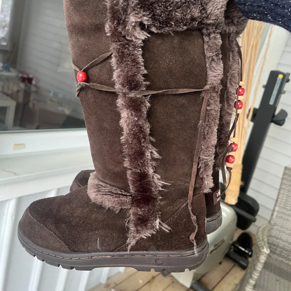 Säljer ett par mysiga bruna UGG-stövlar i mocka med fluffig pälsdetalj. De har dekorativa röda pärlor och snörning på sidan. Perfekta för kalla vinterdagar! 💖Den har mycket små defekter som man kan se på tredje bilden.. Skor.