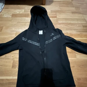 Svart huvtröja från Nike - Säljer en svart huvtröja från Nike med dragkedja och huva. Tröjan har en diskret logga på bröstet och en dekorativ rand över bröstet. Perfekt för en sportig look.