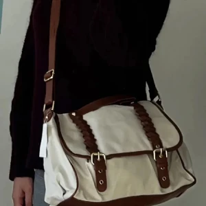 Väska - Säljer en snygg vit axelväska med bruna detaljer. Väskan har en justerbar axelrem och coola flätade detaljer på framsidan. Perfekt för både vardag och fest! Helt ny med lappen kvar. Använd gärna KÖP NU om du vill köpa.