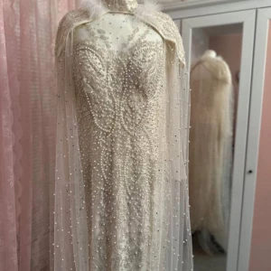 Vit glittrig klänning med pärlor - Säljer en fantastisk vit klänning med glittrande detaljer och pärlor. Den har en elegant cape med fjäderdetaljer vid axlarna. Perfekt för en glamorös fest eller nyårsafton. Klänningen är lång och har en tight passform som framhäver figuren.