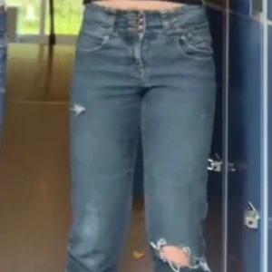 Mörkblå jeans med slitningar - Säljer ett par skitsnygga snygga blå jeans med coola slitningar på benen. Dem är mid waist/low waist och flera knappar i gylfen för en unik look. Perfekta för en avslappnad stil och passar till både vardag och fest. De är i bra skick och redo för nya äventyr!