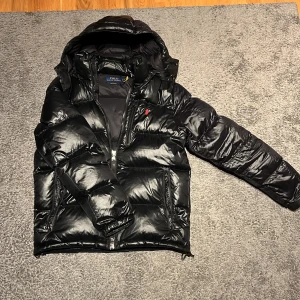 Svart pufferjacka från Polo Ralph Lauren - Säljer en snygg svart pufferjacka från Polo Ralph Lauren i storlek M. Jackan har en glansig finish med en röd broderad logga på bröstet. Den är perfekt för kalla dagar med sin tjocka vaddering och dragkedja framtill samt även en ficka på bröstet. Huvan ger extra skydd mot väder och vind. Perfekt för vintern! 🖤jackan är i jätte bra skick, inga synliga tecken på användning. Kom jätte gärna med prisförslag. Tar även emot byten