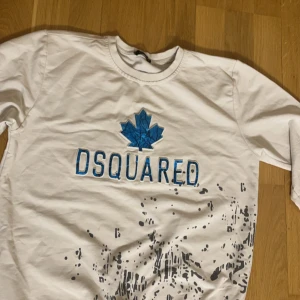 Vit tröja från Dsquared2 - Snygg vit tröja från Dsquared2 med ett blått löv och texten 'DSQUARED' på framsidan. Tröjan har långs ärmar och ett unikt stänkmönster i svart. Perfekt för en avslappnad stil.