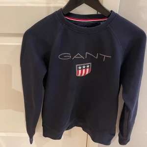 Mörkblå sweatshirt från GANT - Säljer en snygg mörkblå sweatshirt från GANT med deras ikoniska logga och sköld med stjärnor och ränder på bröstet. Tröjan har långa ärmar och är perfekt för en casual look. Den är i bra skick och passar både till vardags och lite mer avslappnade tillfällen.