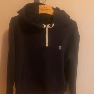 Mörkblå hoodie från Ralph Lauren - Säljer en snygg mörkblå hoodie från Ralph Lauren med en liten vit logga på bröstet. Den har en dragkedja vid halsen och en mysig huva. Perfekt för kyliga dagar eller en avslappnad stil. Passar både till vardags och för en sportig look.