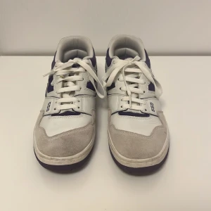 New Balance 550 sneakers - Säljer ett par New Balance 550 sneakers i vitt och lila. Skorna har en snygg retro design med snörning och är i mycket bra skick. Perfekta för både vardag och träning. De har en bekväm passform och är gjorda i en kombination av skinn och syntetmaterial. ÄKTA DISKUTERAS PRISER STORLEK 42.