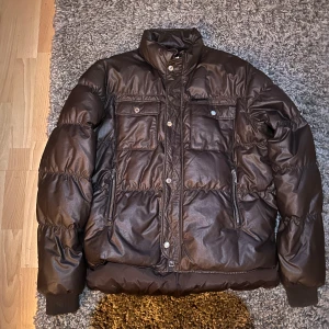 Brun pufferjacka från Dsquared2 - Säljer en snygg brun pufferjacka från Dsquared2. Den har en quiltad design med dragkedja och knappar framtill. Jackan är perfekt för höst och vinter, med långa ärmar och flera praktiska fickor. Den är i mycket bra skick och redo för nya äventyr!