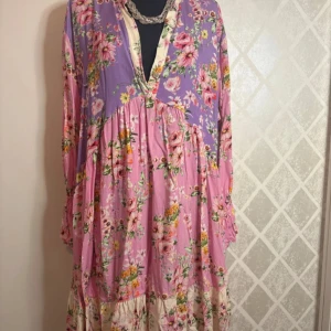 Blommig långklänning i rosa och lila - Charmig långklänning med blommigt mönster i rosa och lila nyanser. Klänningen har långa ärmar och en v-ringad hals. Perfekt för en bohemisk look med volanger och mjukt fall.