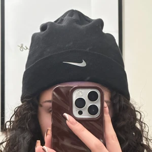 Svart mössa med Nike logotyp - Snygg svart mössa med en silvrig broderad Nike logotyp framtill. Perfekt för att hålla värmen under kyliga dagar. Helt ny - aldrig använd. Super skönt material 