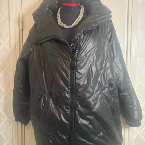 Svart pufferjacka med dragkedja - Snygg svart pufferjacka med stor dragkedja och hög krage. Jackan har en glansig yta och långa ärmar med elastiska muddar. Perfekt för kyliga dagar när du vill hålla dig varm och stilren.