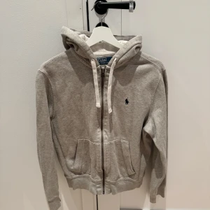 Grå hoodie från Polo Ralph Lauren - Säljer en snygg grå hoodie från Polo Ralph Lauren i storlek S. Den är i bra skick och har en dragkedja framtill samt en klassisk logga på bröstet. Perfekt för höst och vår, och den är supermjuk och bekväm. Passar både till vardags och chillkvällar. 🐴