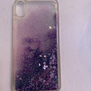 Genomskinligt mobilskal till IPhone xs  med lila glitter och vätska i. - Till Ipone xs. Vet ej var denna är köpt men det finns en liknande på PhoneLife. Skakar man på skalet åker glitteret runt och det är någon slags vätska i. Fint använt skick.