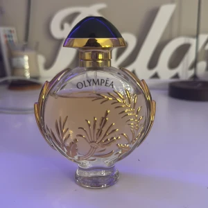Paco Rabanne Olympea intense parfym  - Elegant parfymflaska från Olympea med en unik rund form och dekorativa gulddetaljer. Flaskan har en genomskinlig design som visar den ljusrosa vätskan inuti. Korken är svart med guldfärgad kant, vilket ger en lyxig känsla.