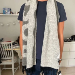 Grå halsduk från BOSS - Snygg och stilren grå halsduk från BOSS med stora bokstäver i mörkare grått. Tillverkad i mjukt material med fransar i ändarna. Perfekt för att hålla dig varm och trendig.