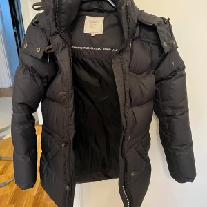Svart dunjacka med huva - Säljer en svart boomerang alexandra down dunjacka i mycket bra skick. Den har en skön huva och stängs med både dragkedja och knappar. Jackan är riktigt varm och mysig! Helt ny!