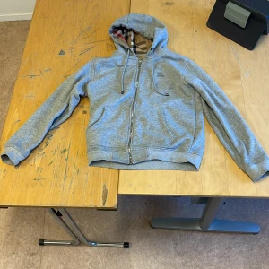 Grå hoodie från Burberry pris kan diskuteras - Säljer en stilren grå hoodie från Burberry med klassiskt rutigt mönster på insidan av huvan. Tröjan har dragkedja framtill och är perfekt för en avslappnad look. Passar bra till både jeans och joggers.