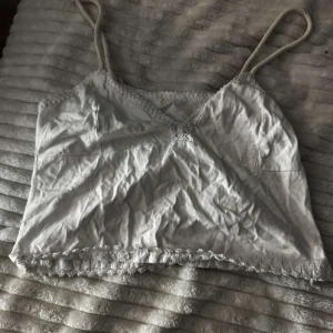 Vit linne med spetsdetaljer - Säljer en söt vit linne med smala axelband och spetsdetaljer vid kanterna. Perfekt för varma sommardagar eller som ett lager under en tröja. Enkel och stilren design. Det är ett linne från Argentina men har inte använt det. Storleken är en xs/s. Vi står för frakt