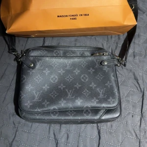 Svart axelväska med monogram - Säljer en snygg svart axelväska med klassiskt monogrammönster. Väskan har en justerbar axelrem och dragkedja för säker förvaring. Perfekt för både vardag och fest! 🖤