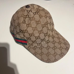 Beige gucci keps 🧢 - Snygg beige gucci  keps med brunt monogrammönster och klassiska ränder i grönt och rött på sidan. Perfekt för en stilren look.🧢