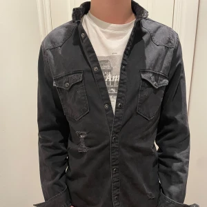 Svart jeansskjorta med slitningar - Säljer en fräsch jeansskjorta/overshirt med snygga slitningar i storlek S. Inga defekter. Skjortan har långa ärmar och knappar framtill samt två bröstfickor med knappstängning. Perfekt för höst och vinter. 