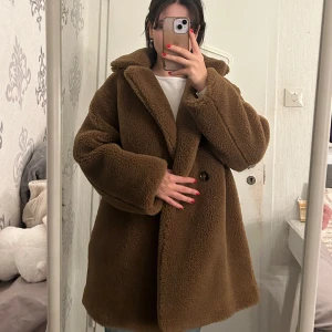 Brun teddyjacka från MANGO - Mysig brun teddyjacka från MNG med stora slag och knappar framtill. Perfekt för kyliga dagar med sin fluffiga och varma design. Jackan har en oversized passform som ger en avslappnad stil.