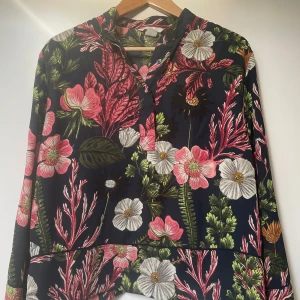 Blommig blus från H&M - Säljer en superfin blommig blus från H&M i storlek M. Den har ett färgglatt mönster med rosa, vita och gröna blommor på en mörkblå bakgrund. Blusen har långa ärmar och knappar framtill. Lyxig kvalitet och look. Otroligt fint skick. Perfekt för alla tillfällen. 