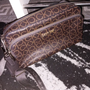 Brun axelväska från Calvin Klein - Snygg brun axelväska från Calvin Klein med deras ikoniska logotypmönster över hela väskan. Den har en justerbar axelrem och en praktisk dragkedja upptill. Perfekt för både vardag och fest!