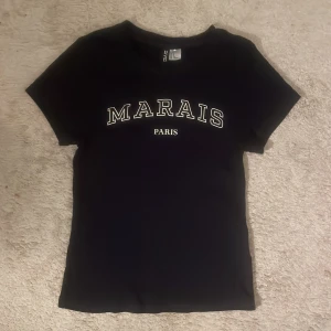 Marinblå t shirt med texten 'Marais Paris' - Säljer en marinblå t-shirt med texten 'Marais Paris' i vitt. Den är i bra skick och har en normal passform. Perfekt för en casual look eller en dag på stan. 🖤