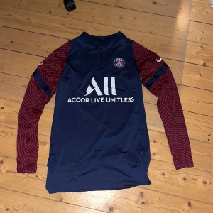 Mörkblå PSG fotbollströja - Säljer en snygg mörkblå PSG fotbollströja från Nike med långa ärmar. Tröjan har röda mönstrade ärmar och ett tryck med 'Accor Live Limitless' på framsidan. Perfekt för fotbollsfans som vill ha något stilrent och bekvämt att bära. Passar både till träning och vardag. Köpt på unisport (äkta)