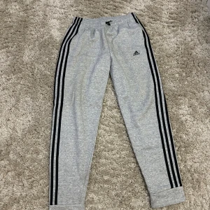 Grå mjukisbyxor från Adidas - Säljer ett par grå mjukisbyxor från Adidas i storlek 164. De har de klassiska tre svarta ränderna längs sidorna och en elastisk midja för extra komfort. Perfekta för träning eller en avslappnad dag hemma. 🖤