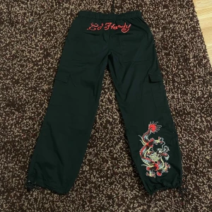 Svarta byxor med drakbroderi från Ed Hardy - Säljer ett par svarta byxor från Ed Hardy med ett coolt drakbroderi i rött och gult på benet. Byxorna har en loose fit och är perfekta för en avslappnad stil. De har elastisk midja och sex fickor. Perfekta för höst och vår! 🌝🐉